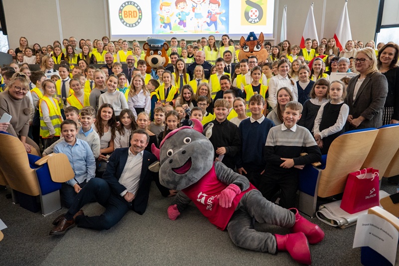 Grupowe zdjęcie dzieci i dorosłych w sali. Na pierwszym planie leży maskotka w czerwonej koszulce, a w tle widać uśmiechniętych uczestników, inne maskotki, ekran z grafiką oraz biało-czerwone flagi fot. FB szkoły